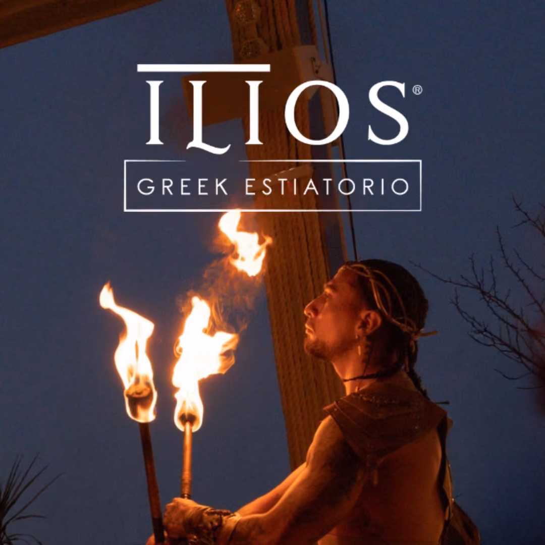 At night, a performer holds two flaming torches and looks up as the ILIOS GREEK ESTIATORIO logo appears above.