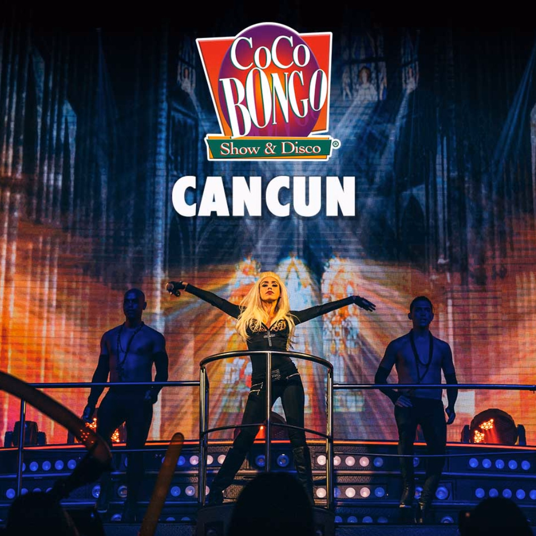 A blonde performer stands with arms outstretched on stage at Coco Bongo Cancun, two male dancers and colorful lights behind.