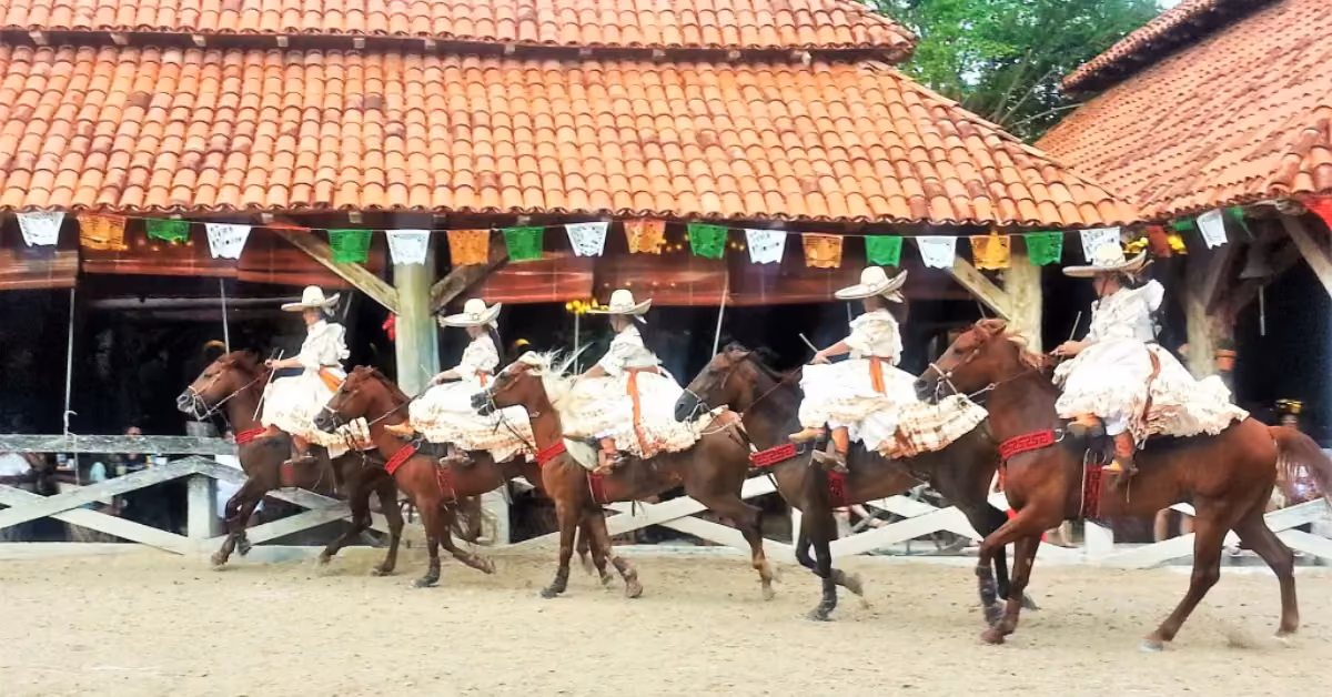 Women in vibrant Mexican dresses and sombreros perform horseback riding at Xcaret, amid colorful banners and rustic buildings.