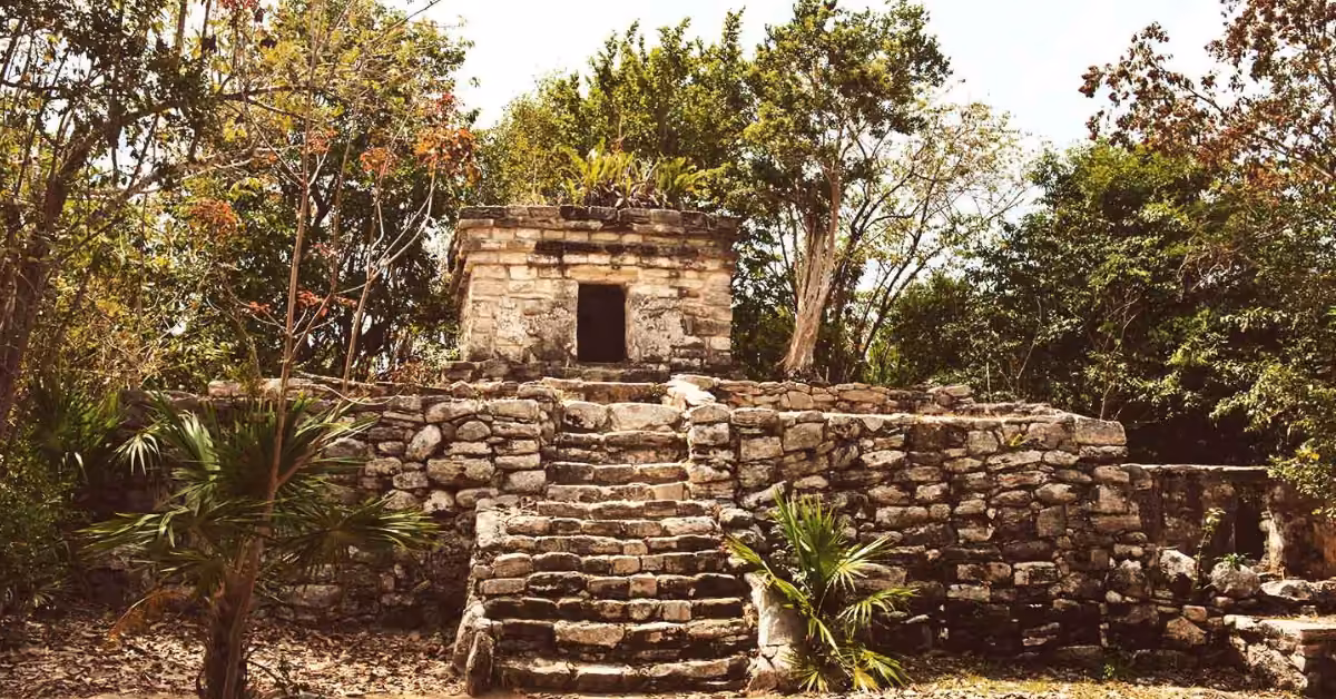 A Mayan stone ruin at Xcaret stands among trees, with a small structure atop a stepped platform and lush greenery all around.
