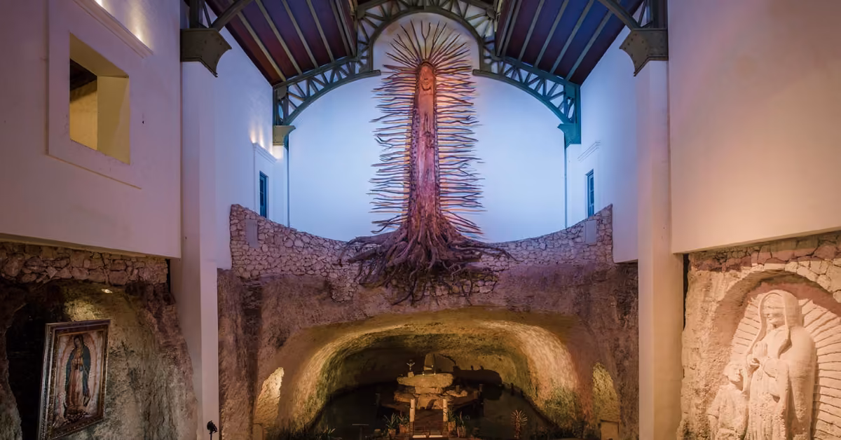 A glowing tree with sprawling roots hovers over a stone altar in a grand, chapel-like hall at Xcaret, surrounded by art.