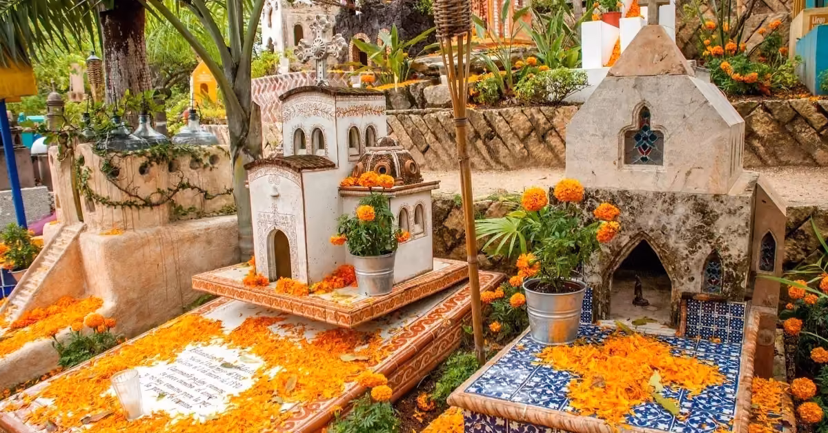 Graves at Xcaret are adorned with orange marigolds, potted plants, and tiny church-like structures in a vibrant outdoor setting.