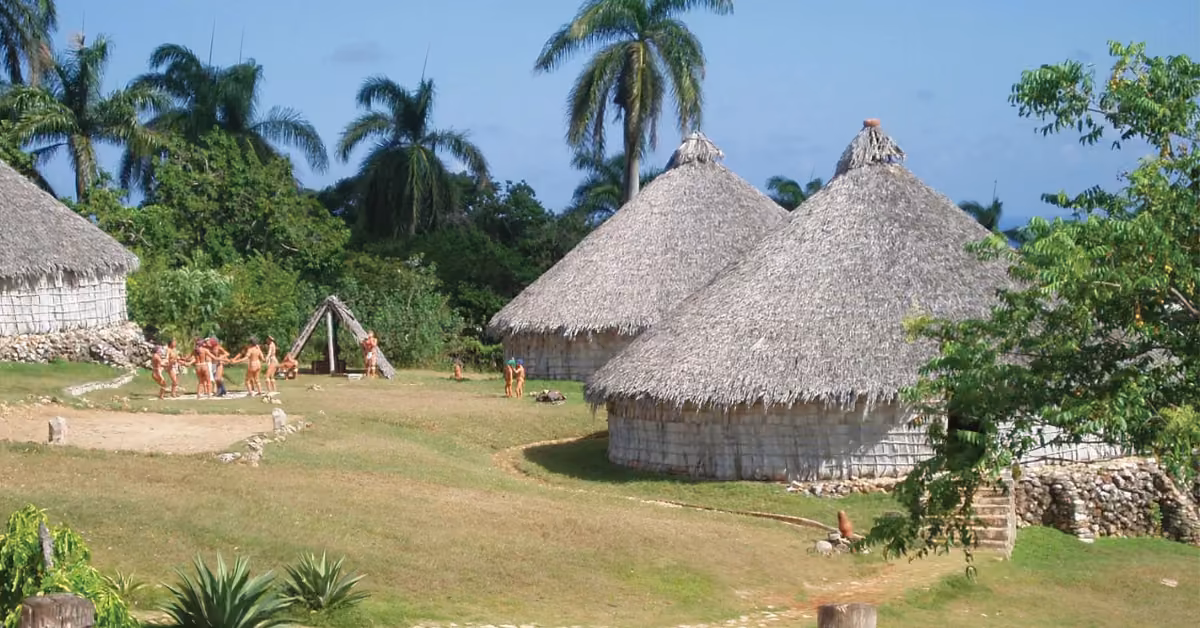 Thatched round huts amid palm trees at Xcaret, with people in traditional dress gathered on the grass nearby.