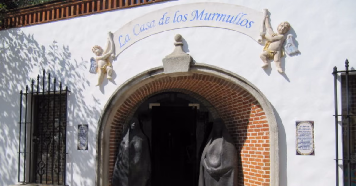 Arched entrance to La Casa de los Murmullos at Xcaret, with two statues, building sign, barred windows, and cherubs.
