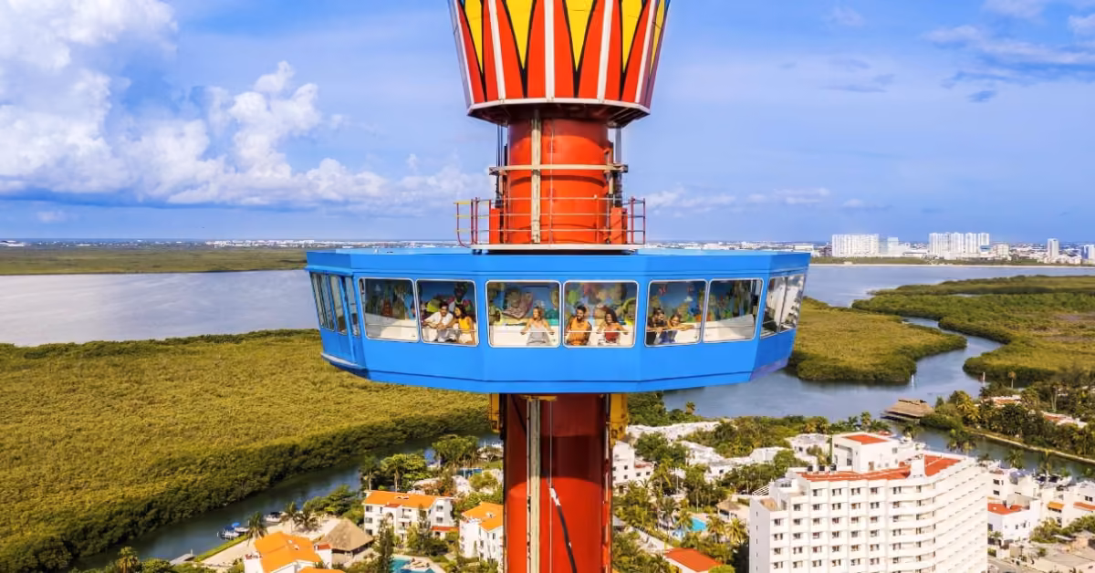 A blue and red observation tower at Xcaret lifts people in a glass cabin over white buildings, mangroves, and sparkling water.