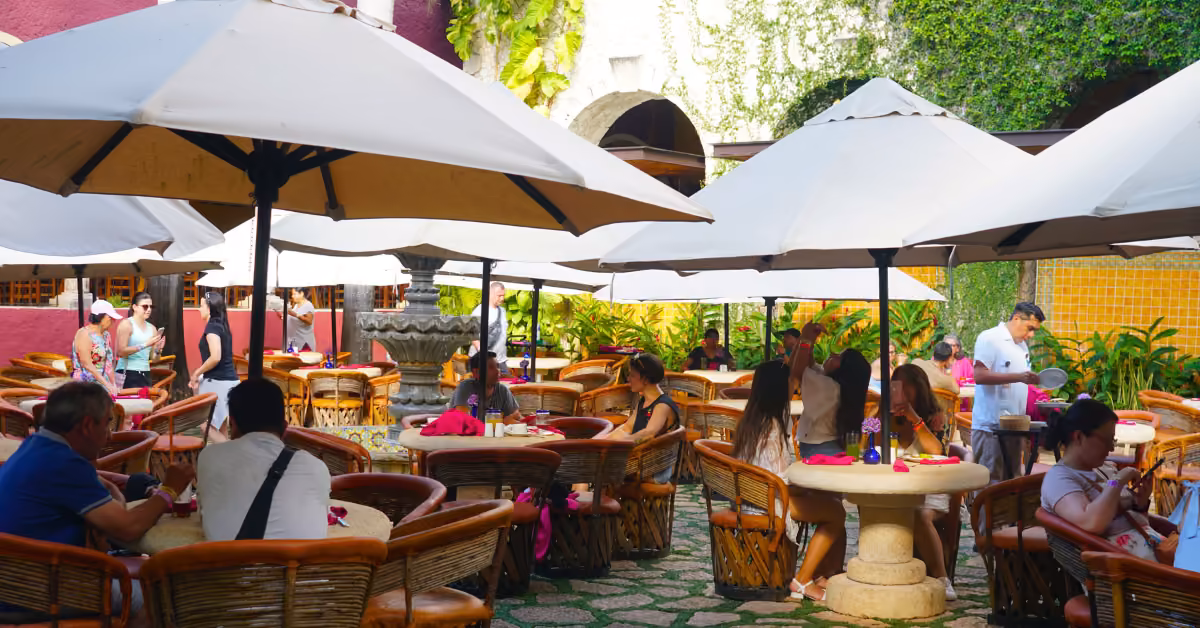 Guests dine and chat at round tables with wicker chairs under white umbrellas, surrounded by greenery and colorful walls.