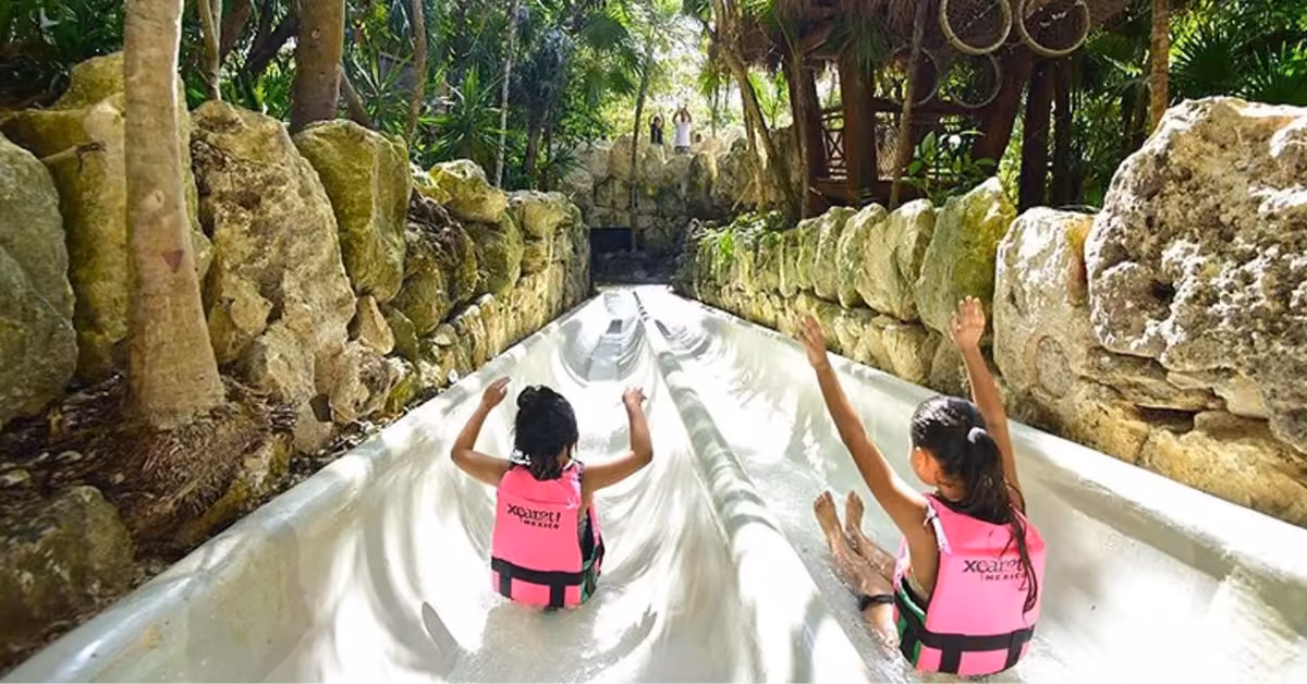 Two kids in pink life jackets slide down a double water slide at Xcaret, surrounded by rocks and lush green trees.