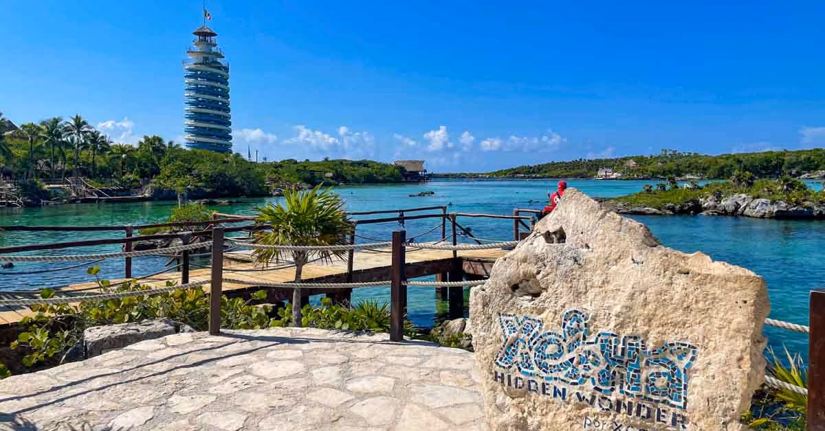 Xel-Ha park with a “Xel-Há Hidden Wonder” stone sign, lagoon, wooden walkways, lush greenery, and tall tower under blue sky.