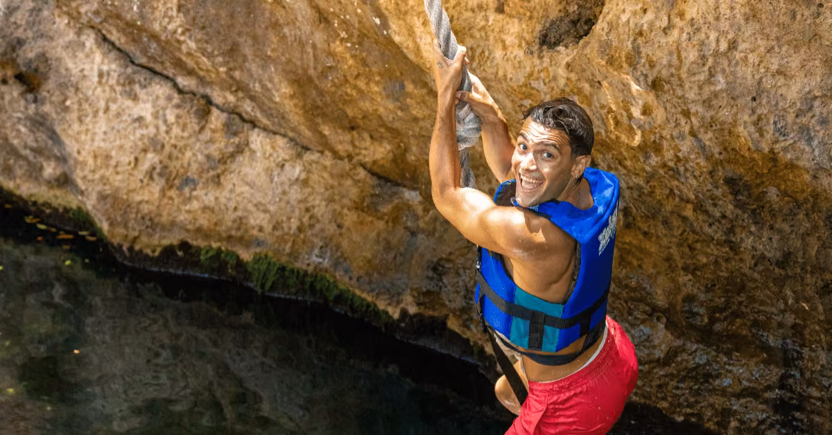 Smiling in a blue life vest and red shorts, a person grabs a rope at Xel-Ha, ready to jump from a rocky ledge into clear water.