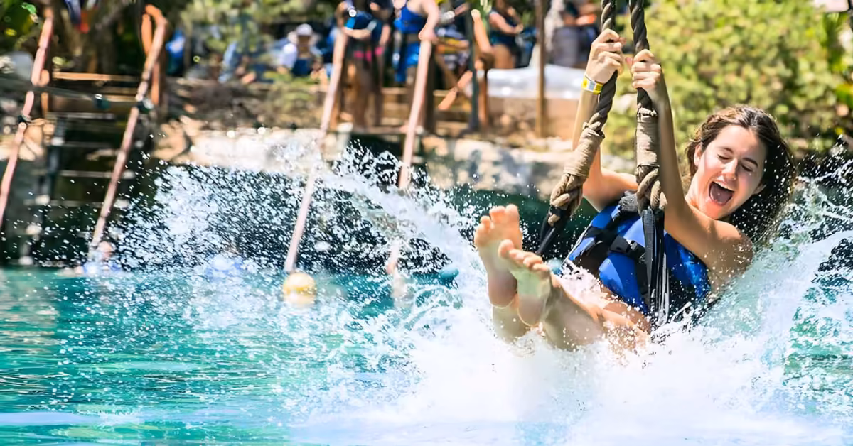 A woman in a life jacket swings on a rope, splashing into bright blue water at Xel-Ha, with people and trees behind her.