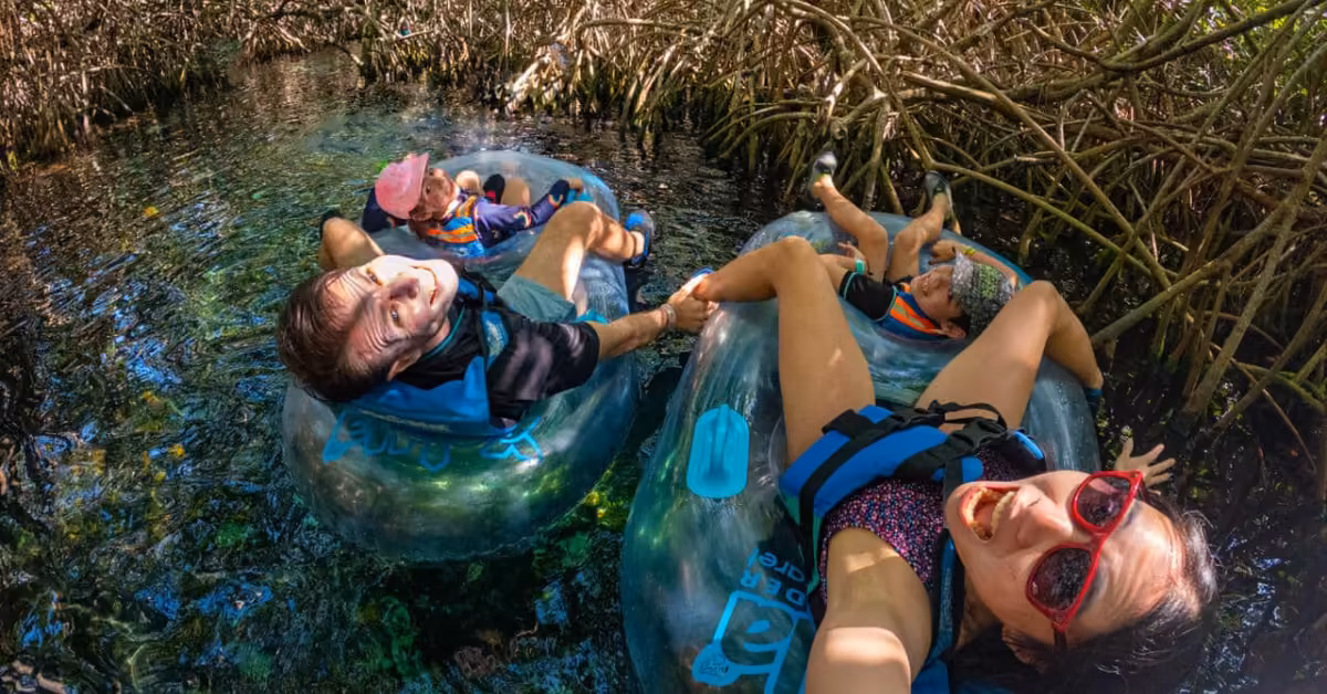 A smiling family in life jackets and sunglasses floats on clear water in inner tubes, enjoying a sunny day among mangroves.