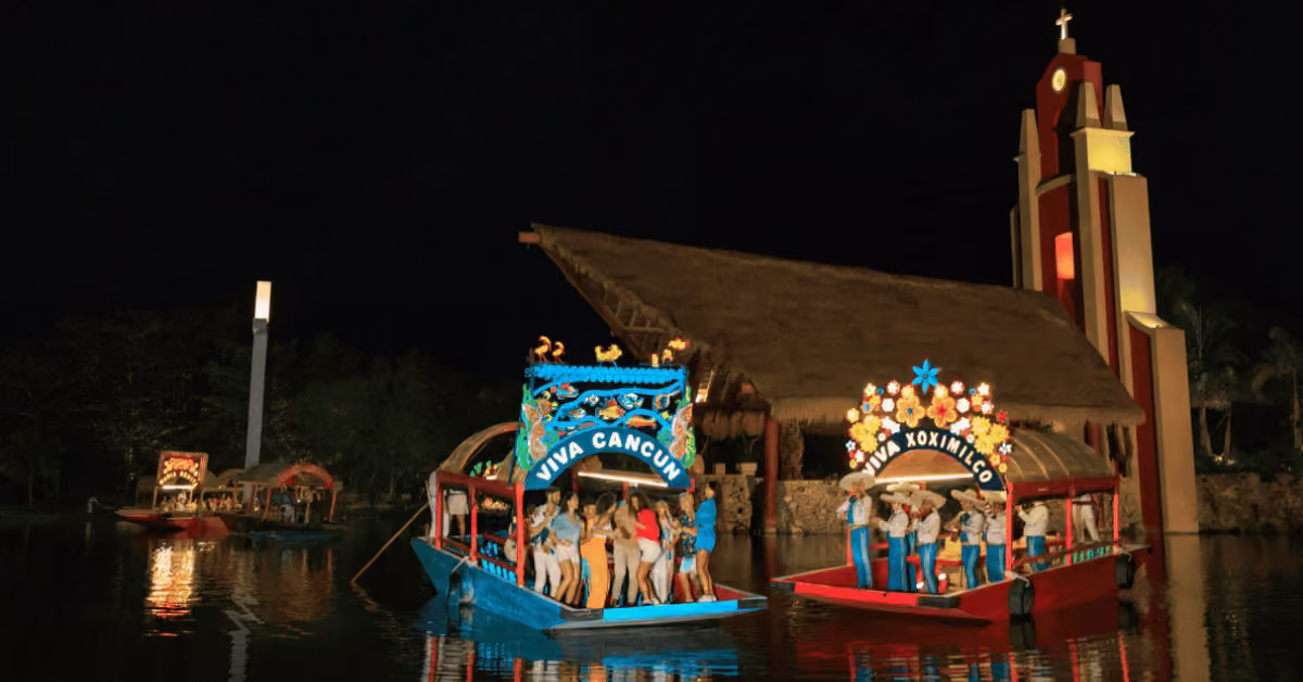 Boats adorned with lights and flowers float at night as people dance nearby, with illuminated buildings in the background.