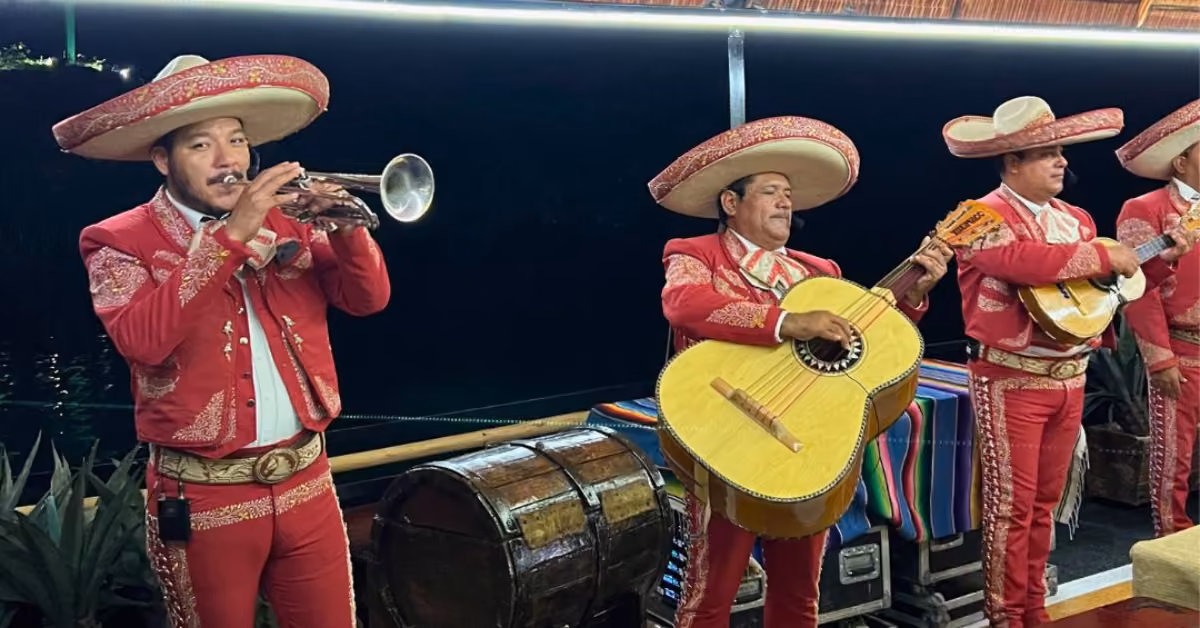 Four mariachi musicians in red and gold perform with trumpet, guitar, and guitarrón beside a drum and colorful blankets.