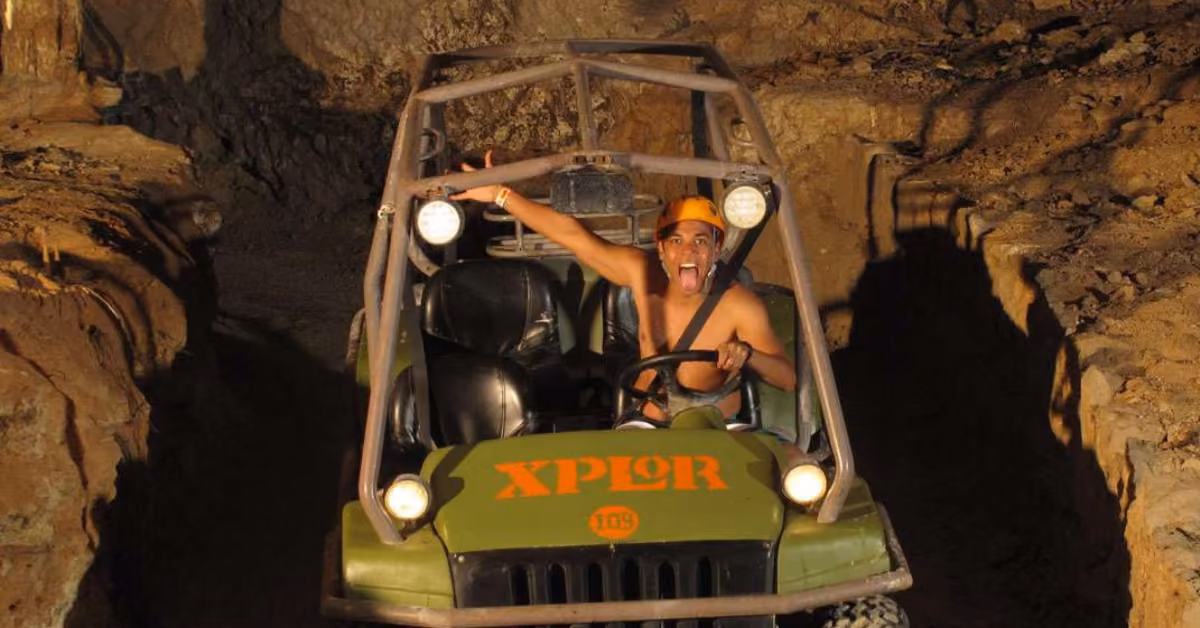 Helmeted driver steers an Xplor off-road vehicle through a rocky cave, arm raised and tongue out as headlights light the walls.