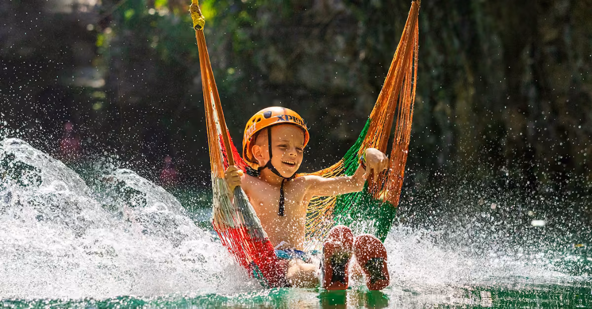 A smiling child in a helmet glides on a colorful hammock over water at Xplor, with lush greenery in the background.