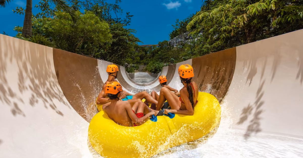 Four people in orange helmets raft down a waterslide at Xplor, surrounded by lush green trees and a bright blue sky.