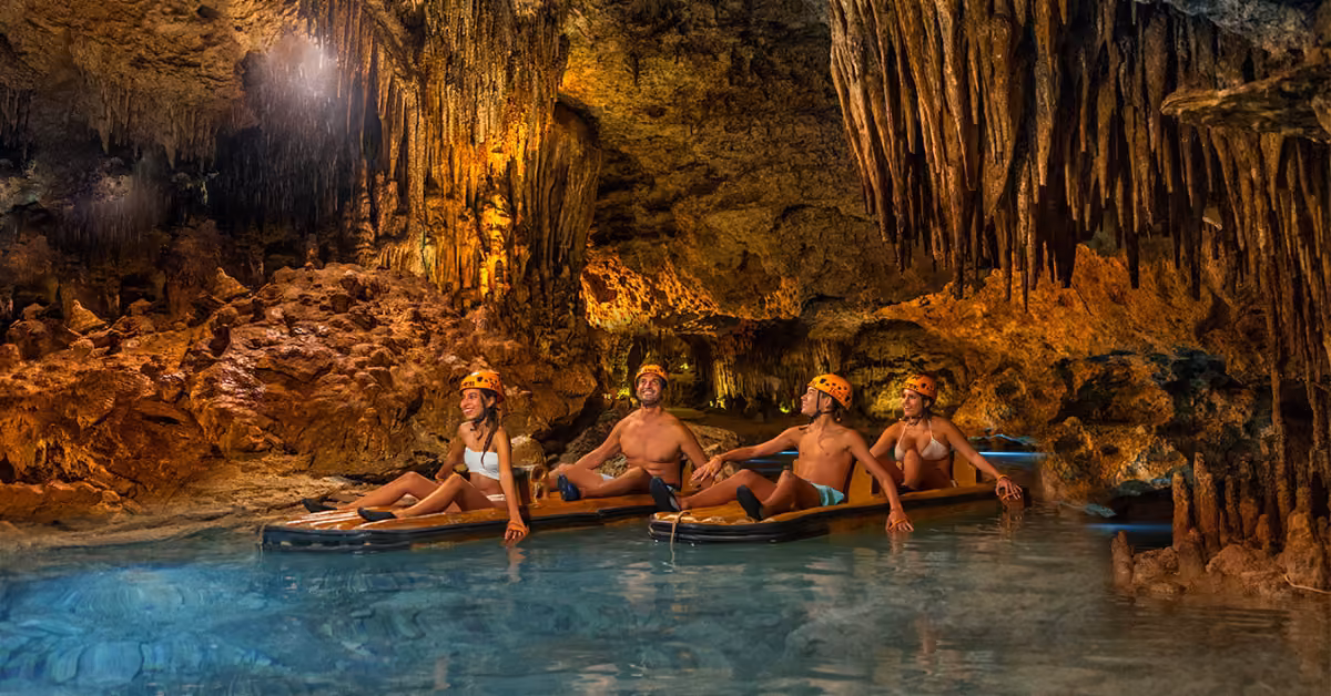 Four people in helmets and swimsuits sit on rafts in a glowing cave at Xplor, with clear water, rocky walls, and stalactites.